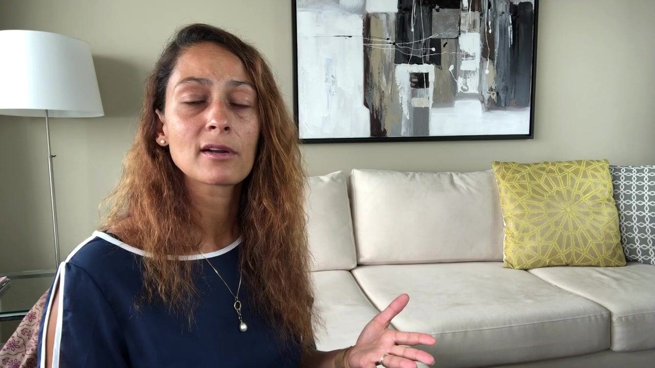 Presenter explaining that drop-off locations and home pickup services exist for sharps disposal, seated in a living room with a beige couch, yellow and gray pillows, a modern painting, and a white floor lamp.