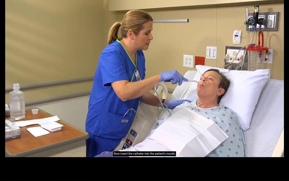 Nurse inserting the Yankauer catheter into the patient's mouth while the patient sits in a semi-Fowler's position, draped, and wearing a nasal cannula.