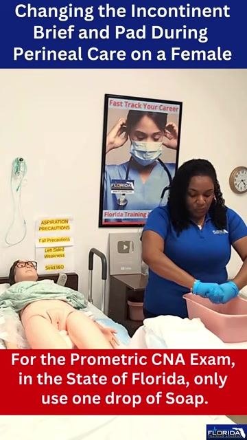 A caregiver in blue gloves removes the soiled incontinent brief from a female mannequin patient lying on a hospital bed, with medical posters, a pink basin, and precaution signs visible in the background.