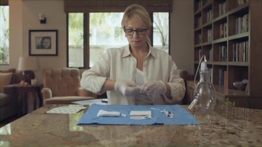 Close-up of gloved hands connecting the drainage line to the catheter valve, seated at a table with a blue sterile drape.