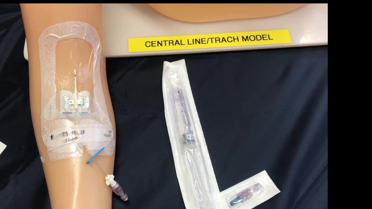 Gloved hands scrubbing the PICC line hub on a training arm, with a saline flush syringe positioned nearby for use