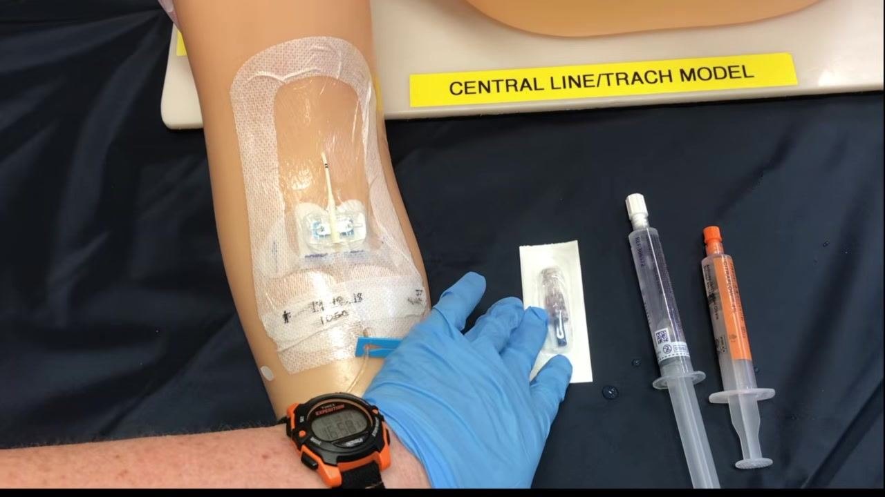 A gloved hand reaching for a new sterile cap in its packaging, with saline and sodium citrate syringes staged nearby on the training arm