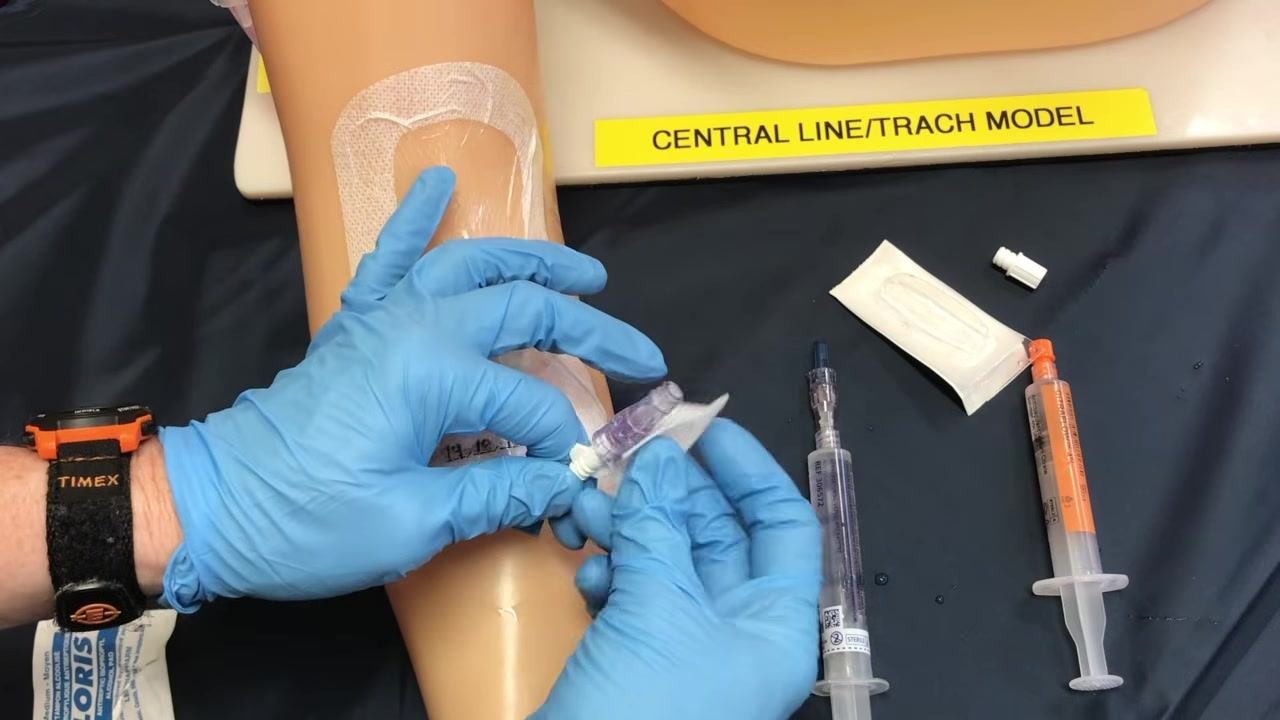 A gloved hand continuing to scrub the PICC line connection site with an alcohol swab before the cap change