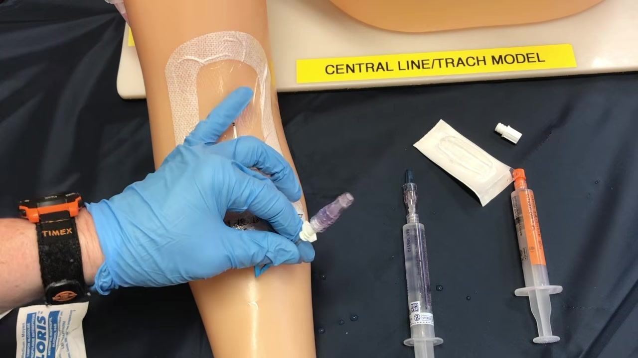 A gloved hand holding the cleansed PICC line connection site on the training arm while it air dries, with staged supplies including a saline syringe, a sodium citrate or heparin syringe, a packaged sterile cap, and the removed cap