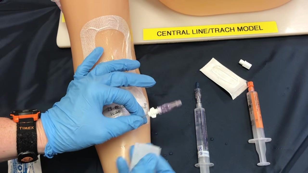 A gloved hand holding the new sterile cap and preparing to attach the saline flush syringe for priming