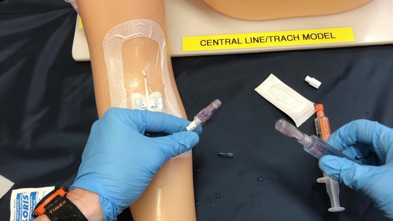 A gloved clinician holding a PICC line extension set and a syringe on a training arm, with supplies arranged on a sterile field
