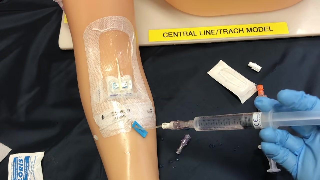 A gloved clinician administering medication through a syringe attached to the PICC line on the training arm
