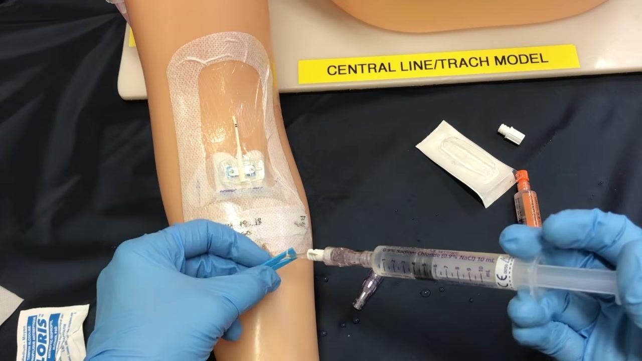A gloved clinician clamping the PICC line while administering the last portion of fluid with a syringe