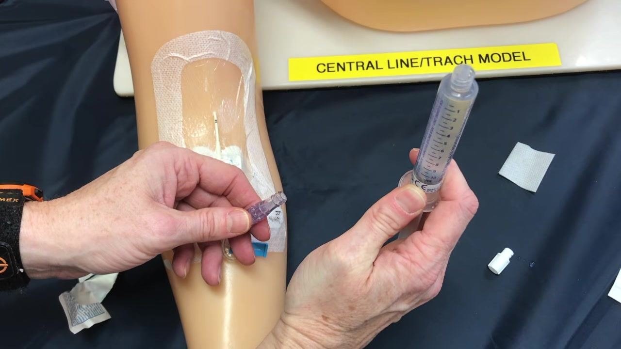Gloved hands holding the PICC line hub and a saline flush syringe, preparing to connect them, with the clamp and supplies visible on the field