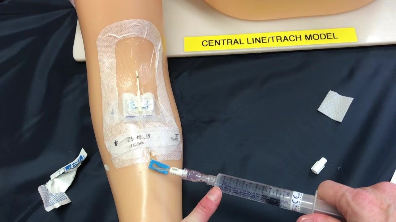 A gloved hand injecting saline into a PICC line on a training arm secured with a transparent dressing, with supplies arranged nearby