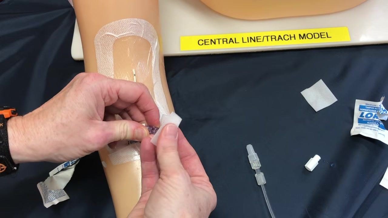 The saline syringe disconnected from the PICC line hub, which remains clamped, with supplies arranged around the training arm