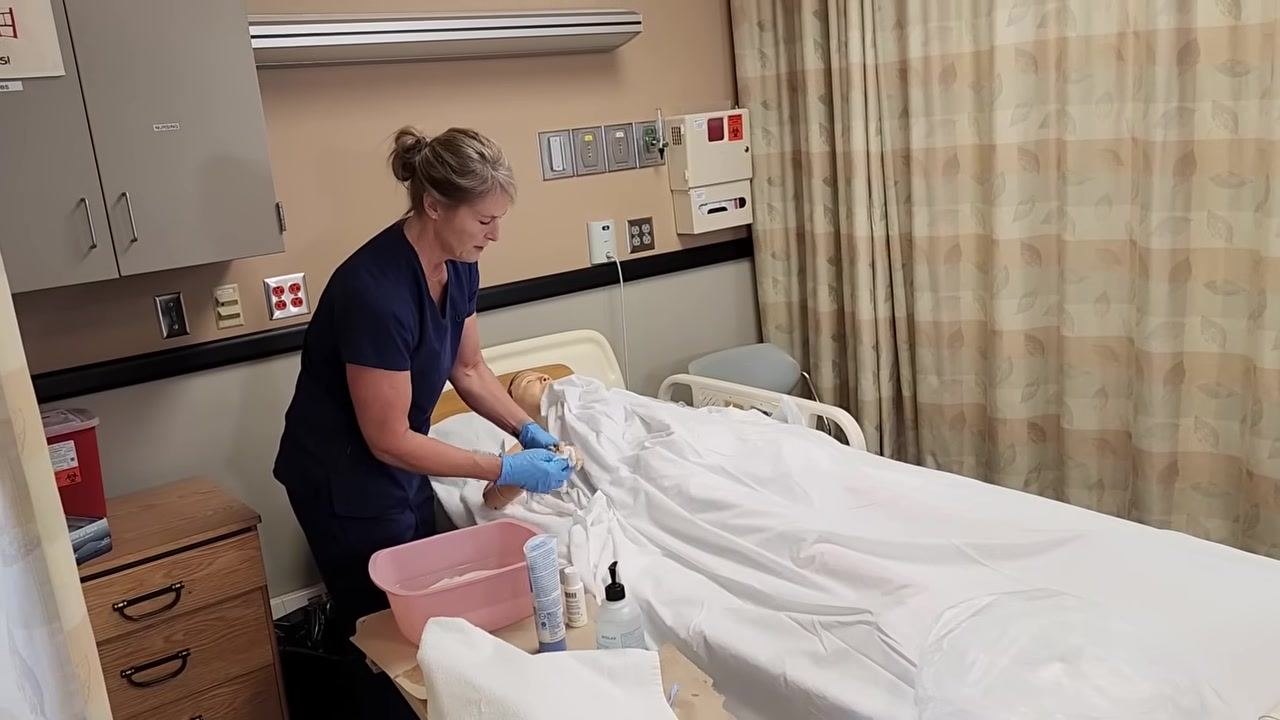 Nurse in blue scrubs washing the mannequin patient's arm with a soapy washcloth, towel underneath to protect the bed.