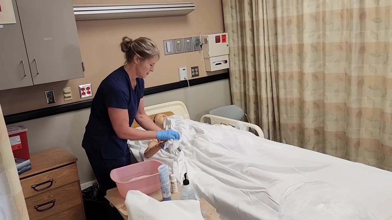 Nurse in blue scrubs, wearing gloves, washing the mannequin patient's chest under the linens, holding the sheet up to maintain warmth and privacy. Pink basin and hygiene supplies are on the bedside table.