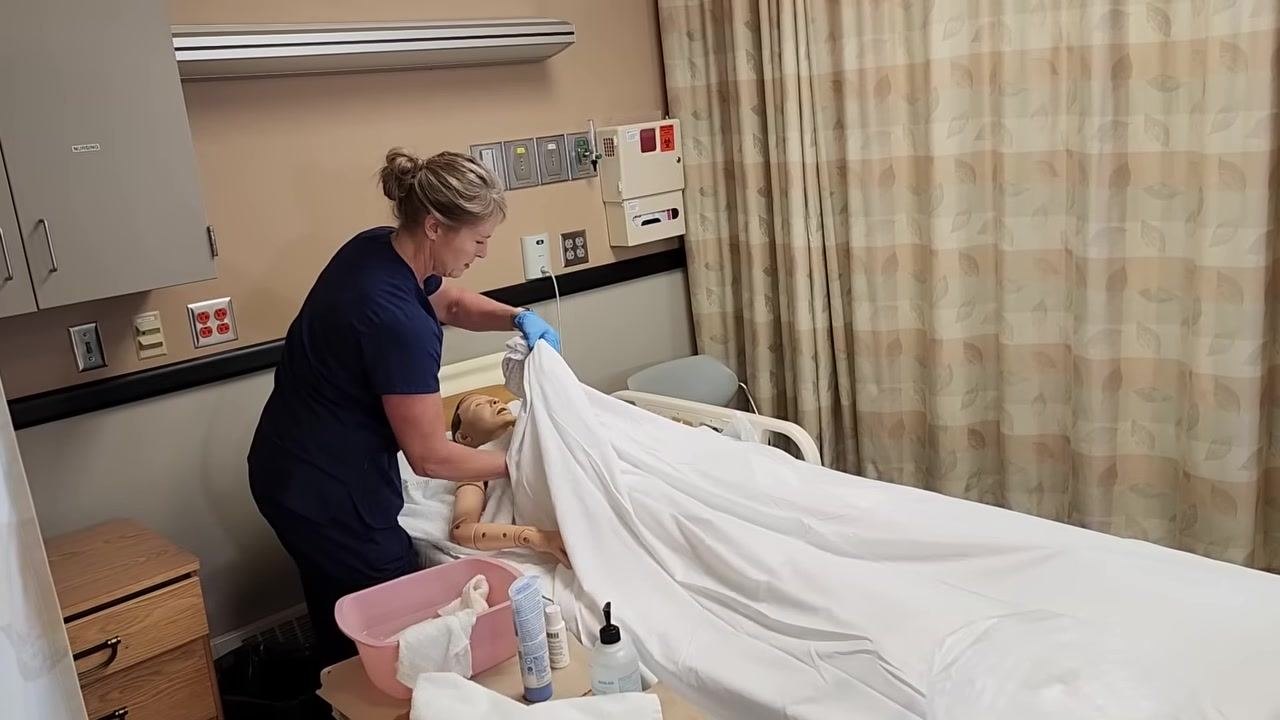 Nurse in blue scrubs patting the mannequin patient's chest dry with a towel, holding up the linens for privacy and warmth.