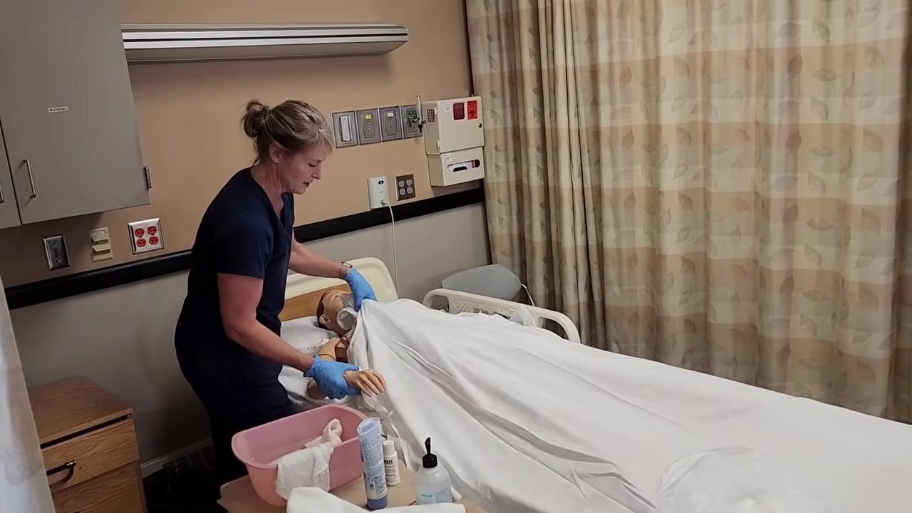 Nurse in blue scrubs patting the mannequin patient's arm dry with a towel, ensuring all areas are dry. Supplies and pink basin visible on the bedside table.