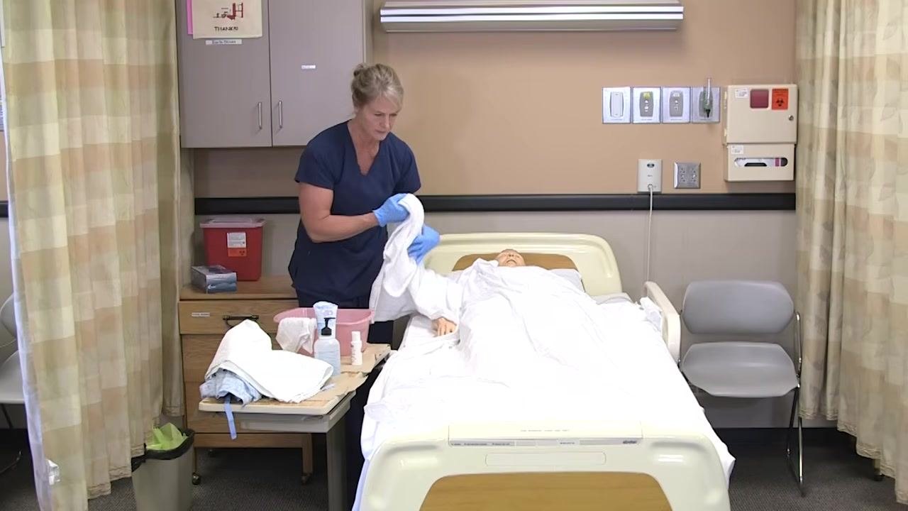 Nurse in blue scrubs preparing to wash the lower body, holding a towel and standing beside the mannequin patient, who is covered except for the arm.