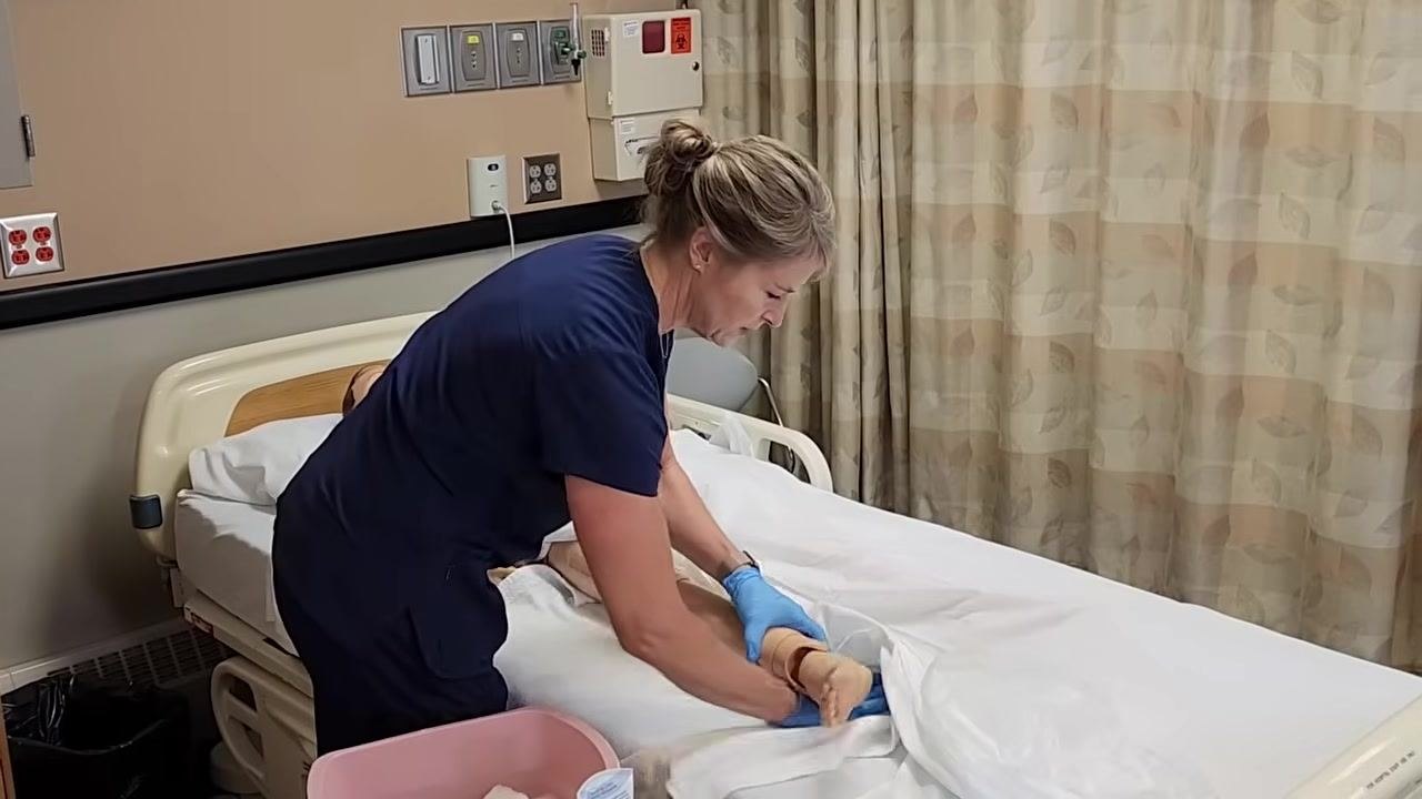 A healthcare worker in navy scrubs and blue gloves is lifting a mannequin's leg on a hospital bed, preparing to wash underneath. A pink basin with supplies is on the bedside table. The environment is a clinical room with privacy curtains and medical wall panels.