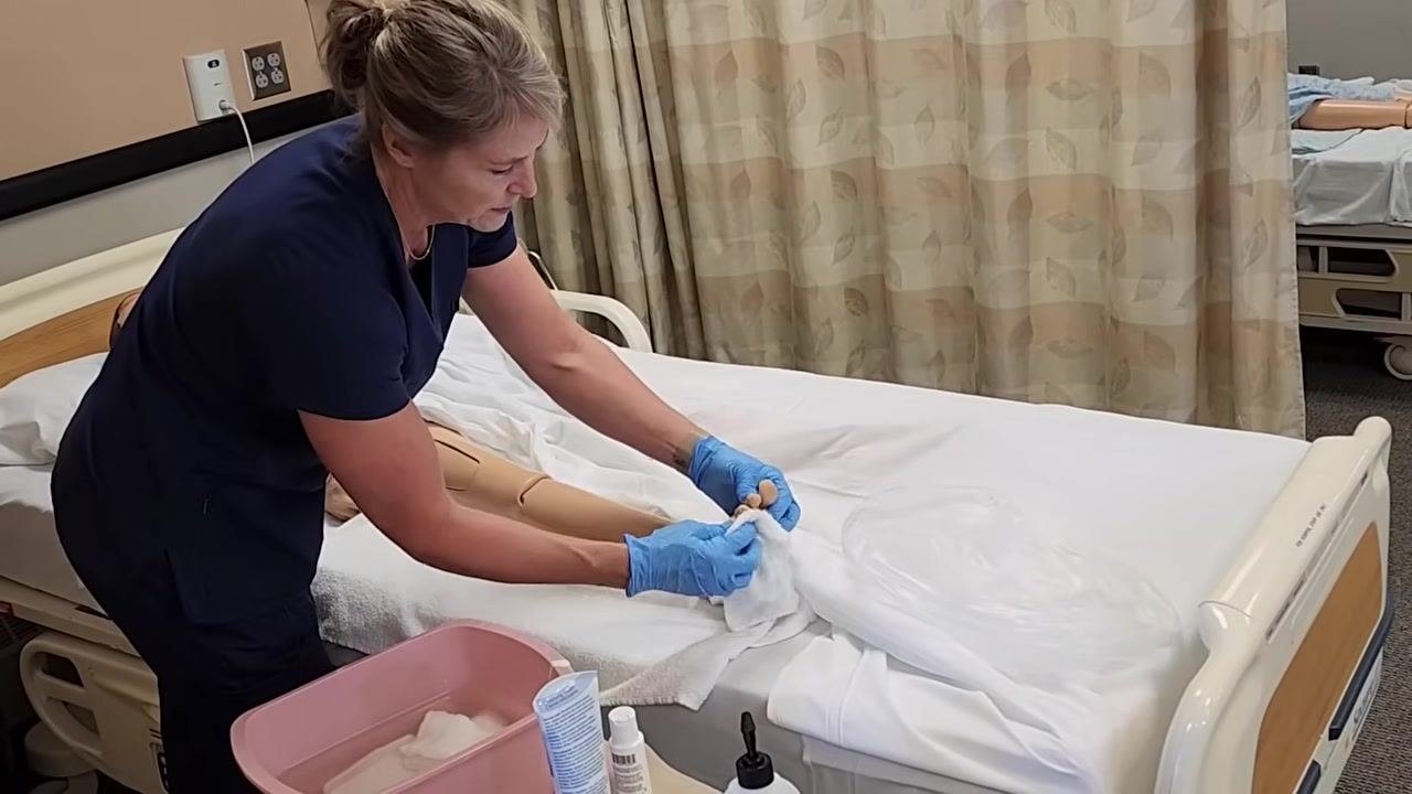 The healthcare worker is holding a soapy washcloth and demonstrating cleaning the mannequin's leg, with the leg resting on a towel. The pink basin and hygiene supplies are visible.