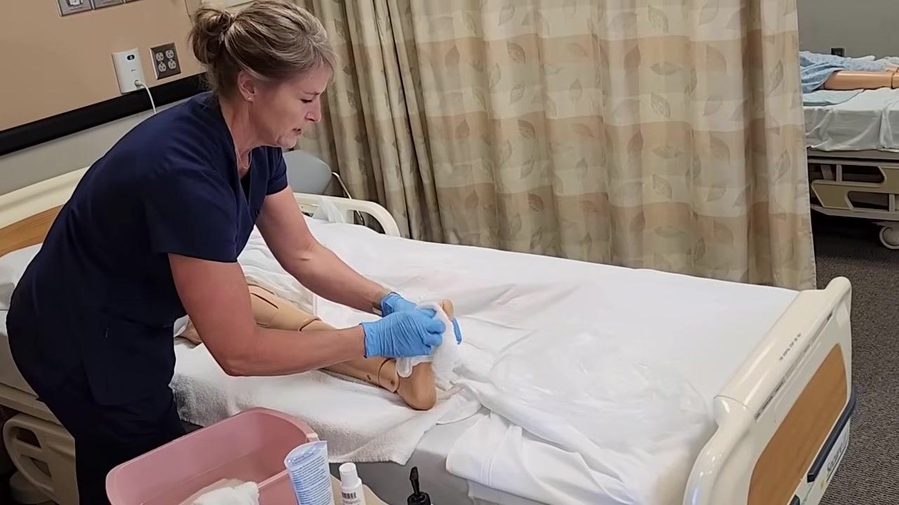 The healthcare worker is rinsing the mannequin's foot and toes with a clean washcloth, focusing on between the toes. The leg is supported on a towel, and supplies are nearby.