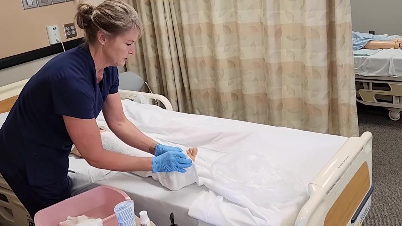 The healthcare worker is drying the mannequin's foot and toes with a towel, ensuring all moisture is removed from skin creases.