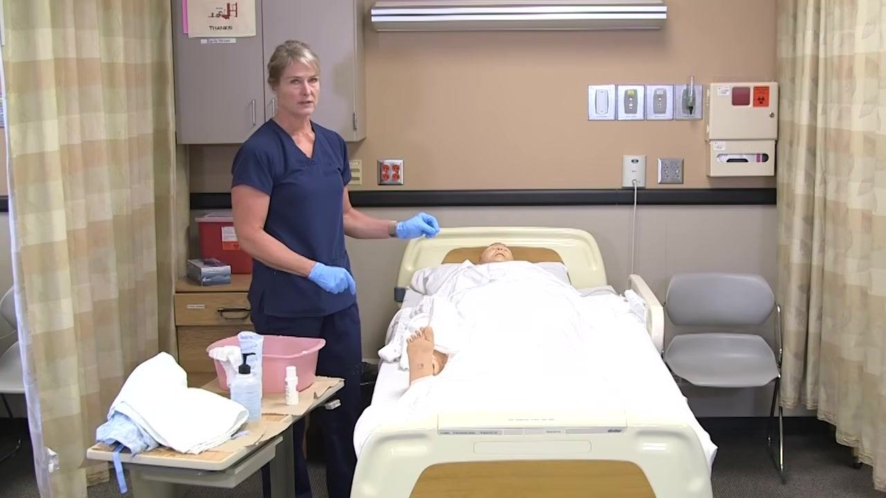 A healthcare worker in navy scrubs and blue gloves stands beside a hospital bed with a mannequin patient. A pink basin, towels, and hygiene supplies rest on a bedside table. The clinical room has privacy curtains, medical wall panels, and a sharps container.