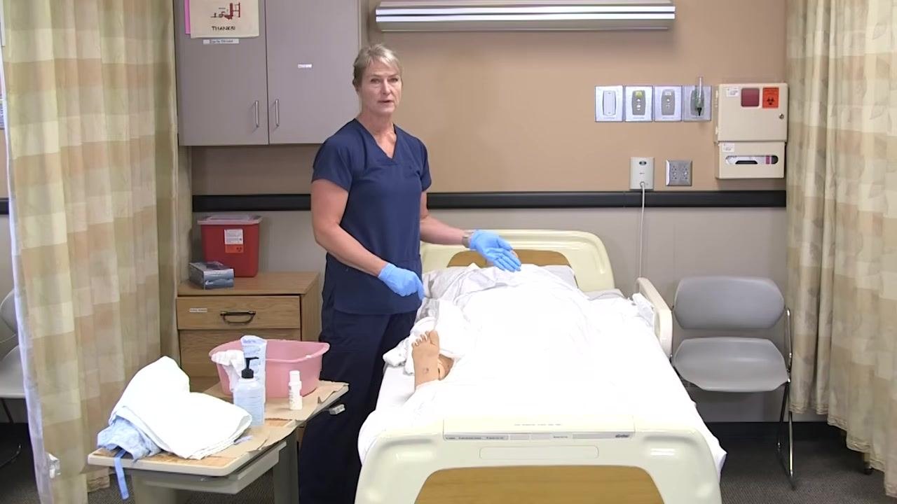 The healthcare worker gestures toward the mannequin patient, indicating the next step. The supplies remain organized on the bedside table.