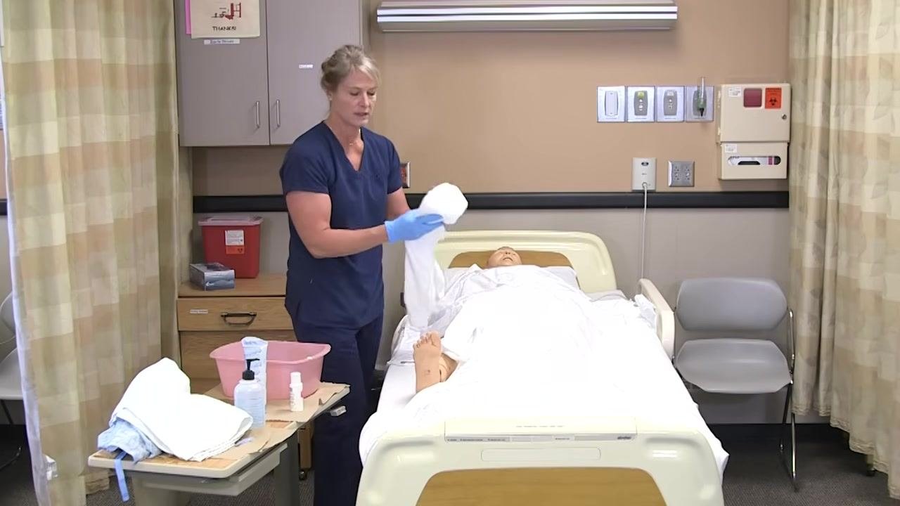 The healthcare worker is holding a clean towel and preparing to change gloves, standing beside the bed and supplies.