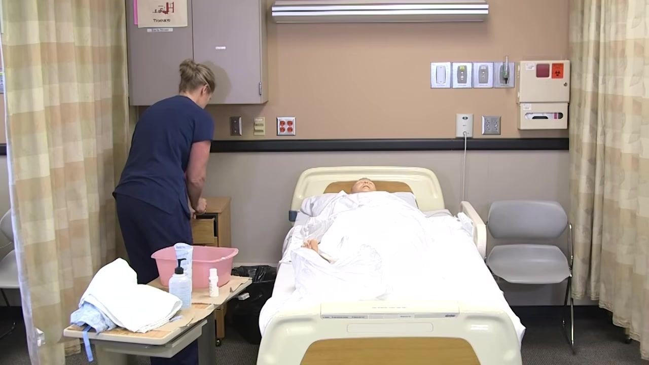 The healthcare worker is turned toward the bedside cabinet, disposing of linens. The mannequin remains covered on the bed.