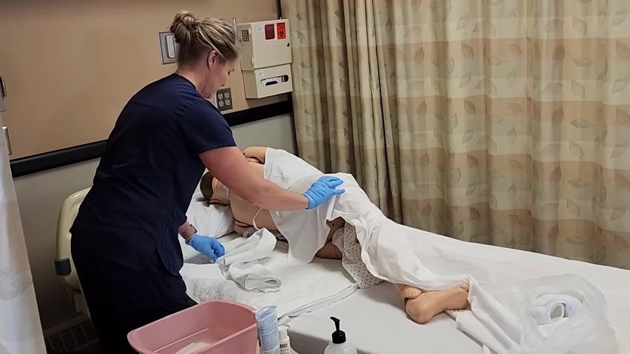 The healthcare worker is ensuring the mannequin patient is properly positioned on their side, with the back exposed and the rest of the body covered. Supplies are ready on the bedside table.