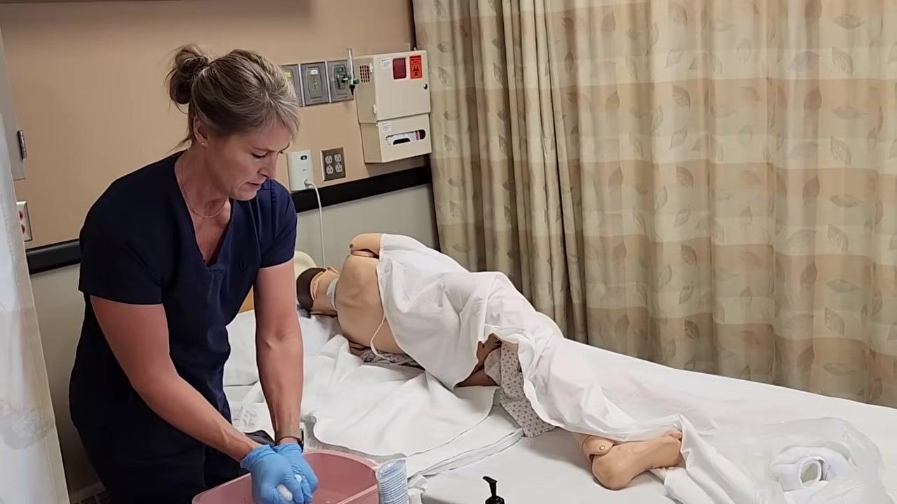 A healthcare worker in navy scrubs and blue gloves prepares a clean washcloth at the bedside. The mannequin patient is lying on their side, covered with a sheet except for the back. Supplies including a pink basin, hygiene products, and towels are organized on the bedside table. The clinical room has privacy curtains and medical wall panels.