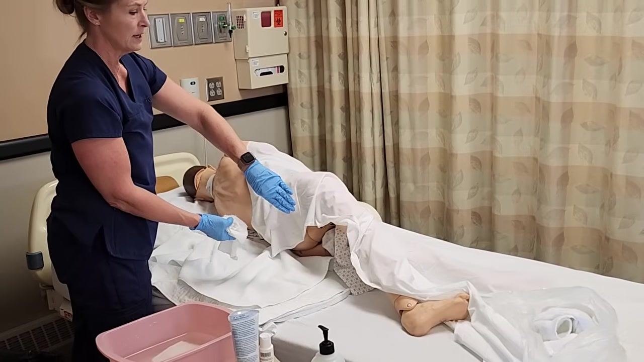 The healthcare worker is gently washing the patient's back with a washcloth, lifting the sheet just enough to access the area while maintaining privacy.