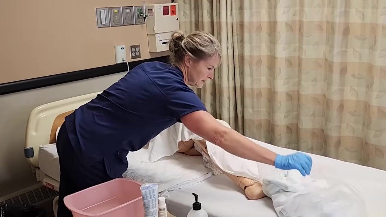 The healthcare worker is rinsing the patient's back and possibly the back of the legs, maintaining proper technique and coverage.