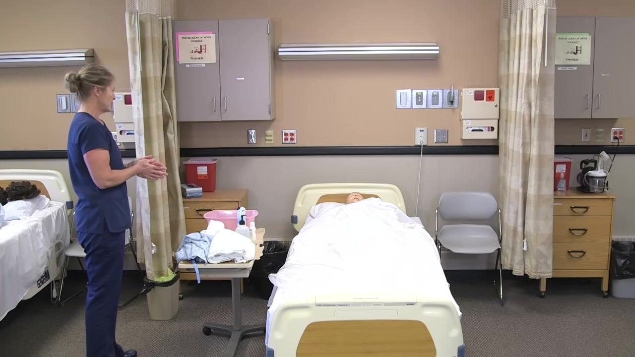 Nurse in blue scrubs adjusting the bed controls, raising the bed with the mannequin patient lying under a white sheet.