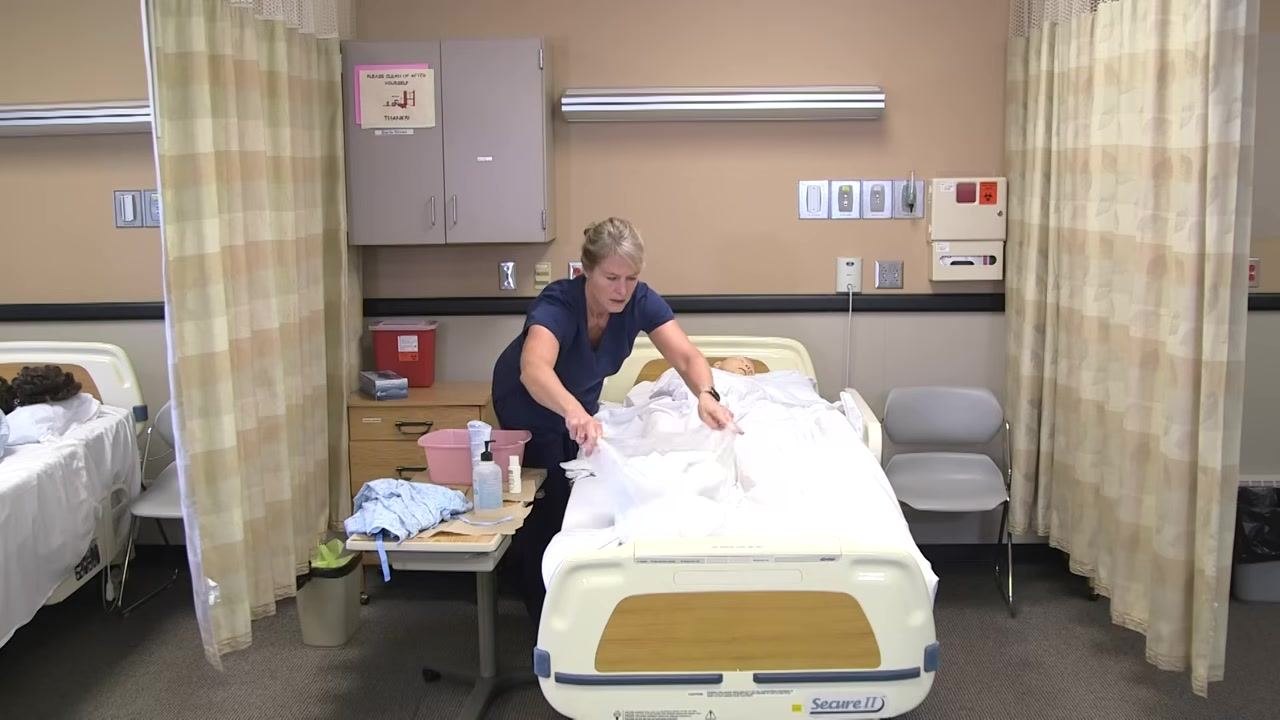 A healthcare worker in navy scrubs is at the bedside of a patient mannequin, preparing linens and a gown. The environment is a clinical room with privacy curtains, medical supplies on a rolling table, and cabinets on the wall.