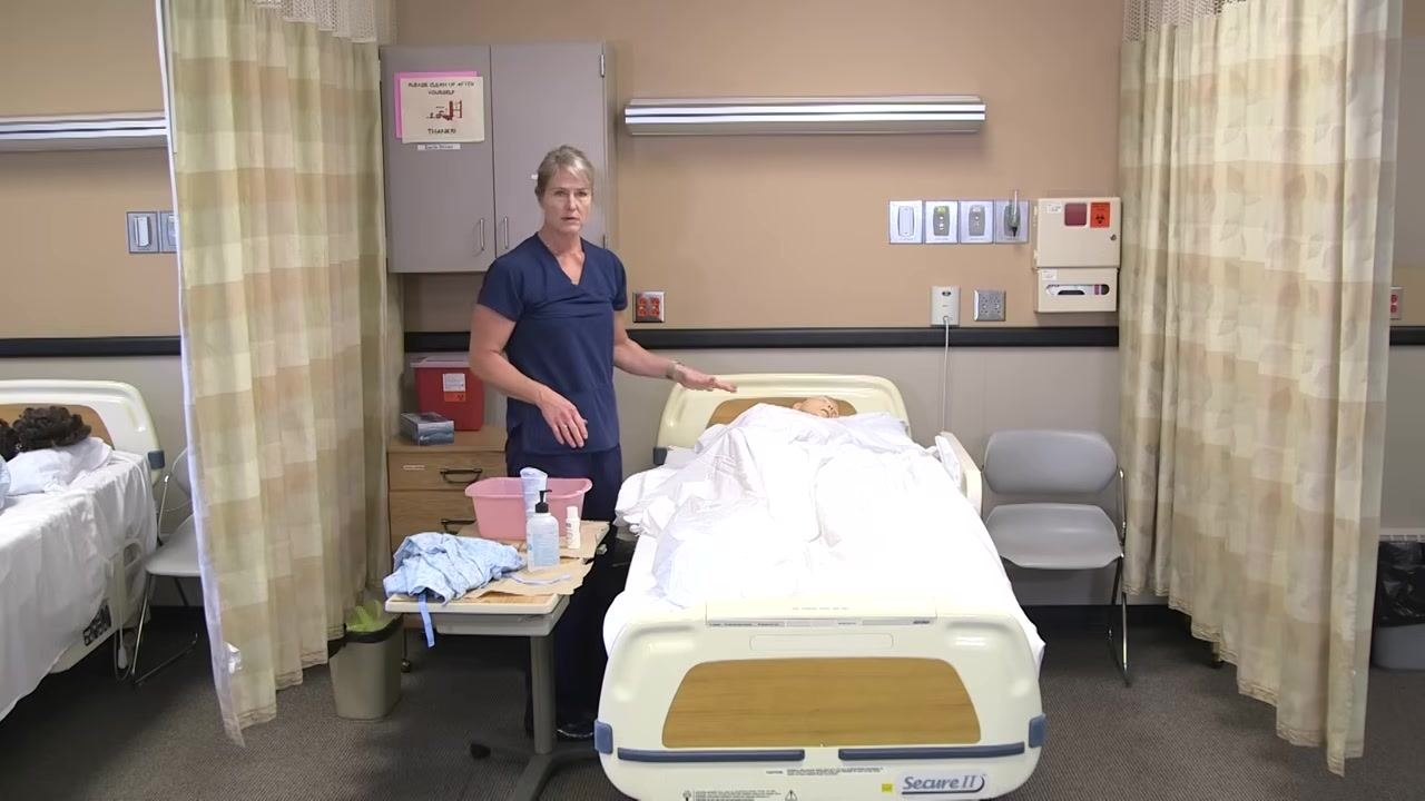 The healthcare worker is standing at the bedside, gesturing toward the patient mannequin, with dirty linens visible on the bed and a pile of supplies on the rolling table.