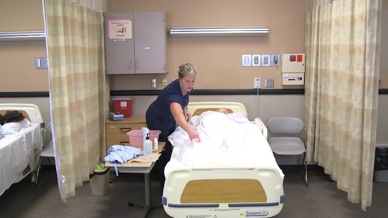 The healthcare worker is removing dirty linens from the bed, preparing to dispose of them. The clinical setting and supplies remain visible.