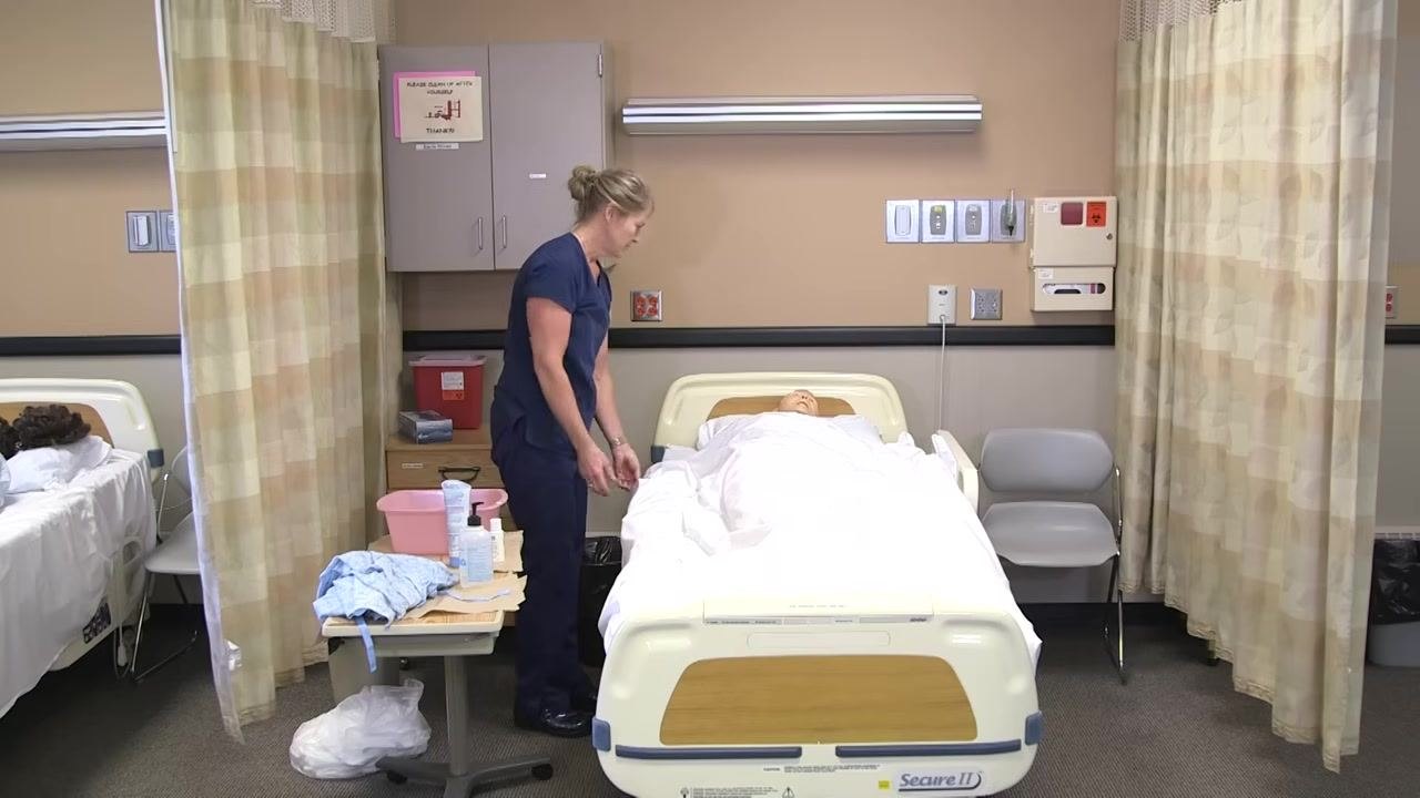 The healthcare worker stands at the bedside, checking on the patient mannequin's comfort. The bed is made, and supplies are organized on the table.