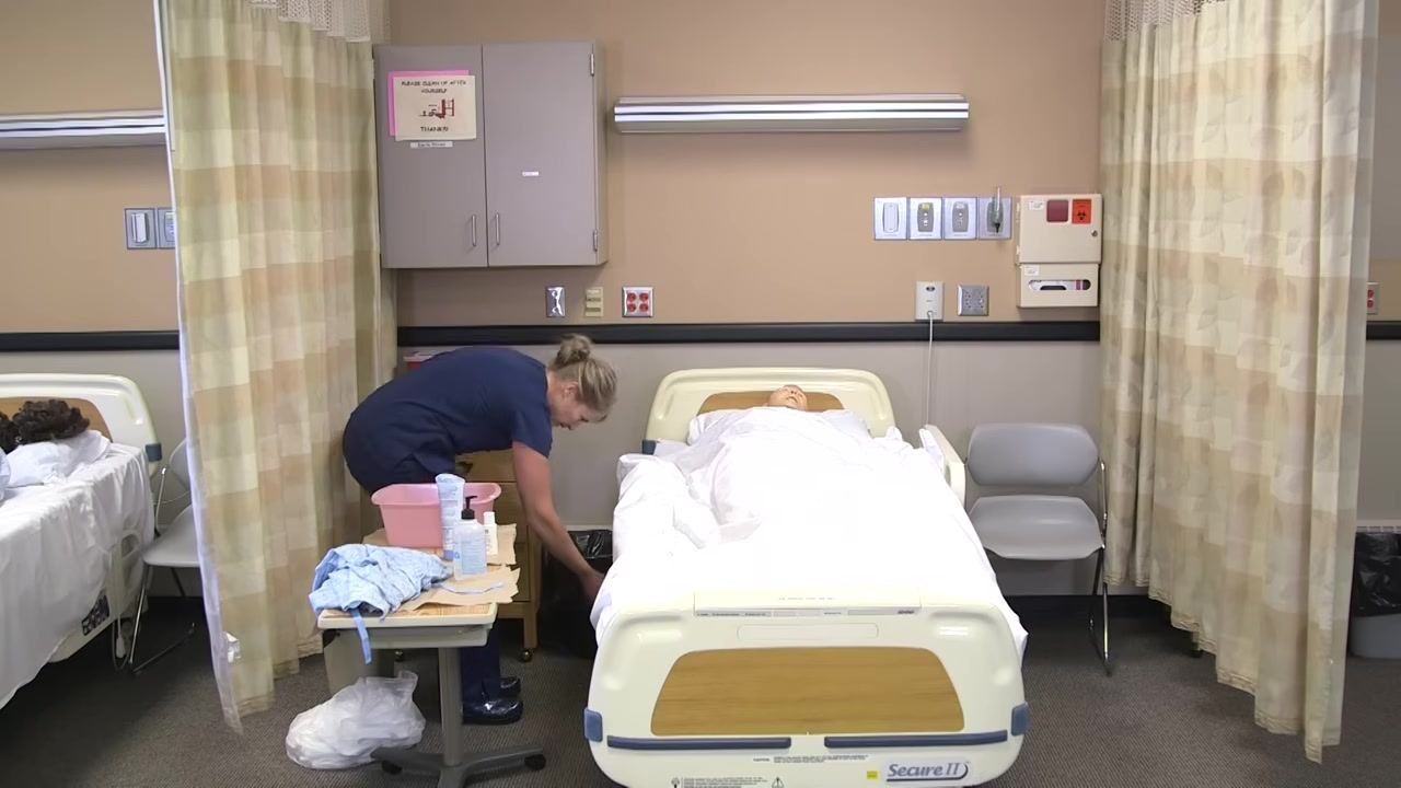The healthcare worker bends down to dispose of items, maintaining a clean environment. The patient mannequin remains in bed, covered.