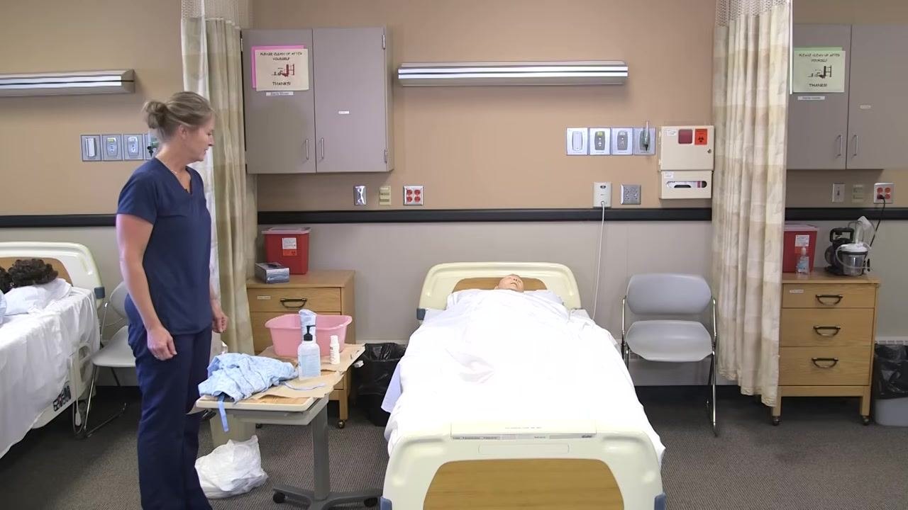 The healthcare worker stands near the bed, with the patient mannequin resting comfortably and the area tidy. The linen hamper is visible.