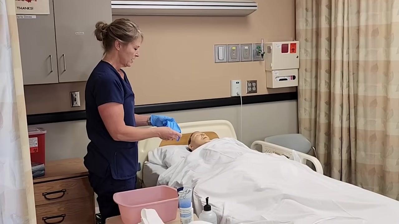 Nurse in blue scrubs putting on blue gloves, standing next to the bed and supplies.