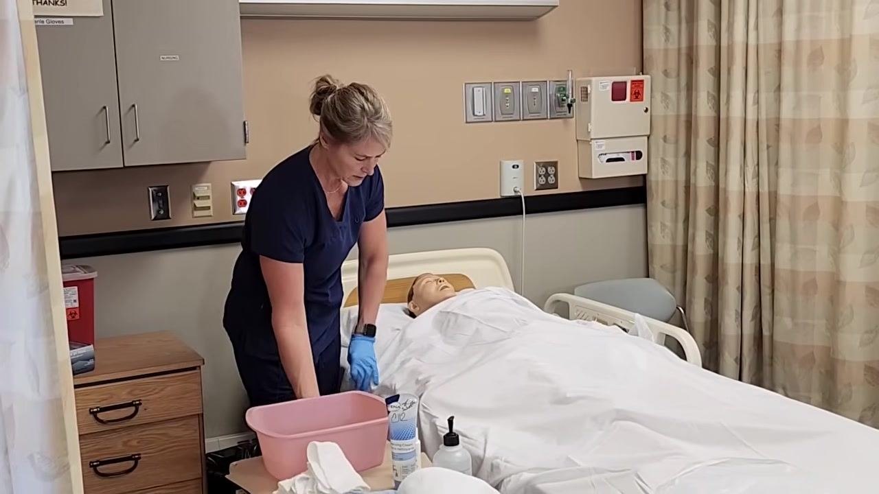 Nurse in blue scrubs, wearing gloves, standing beside a hospital bed with a mannequin patient. She is preparing to wet a washcloth in a pink basin on the bedside table, which also holds hygiene supplies and linens.