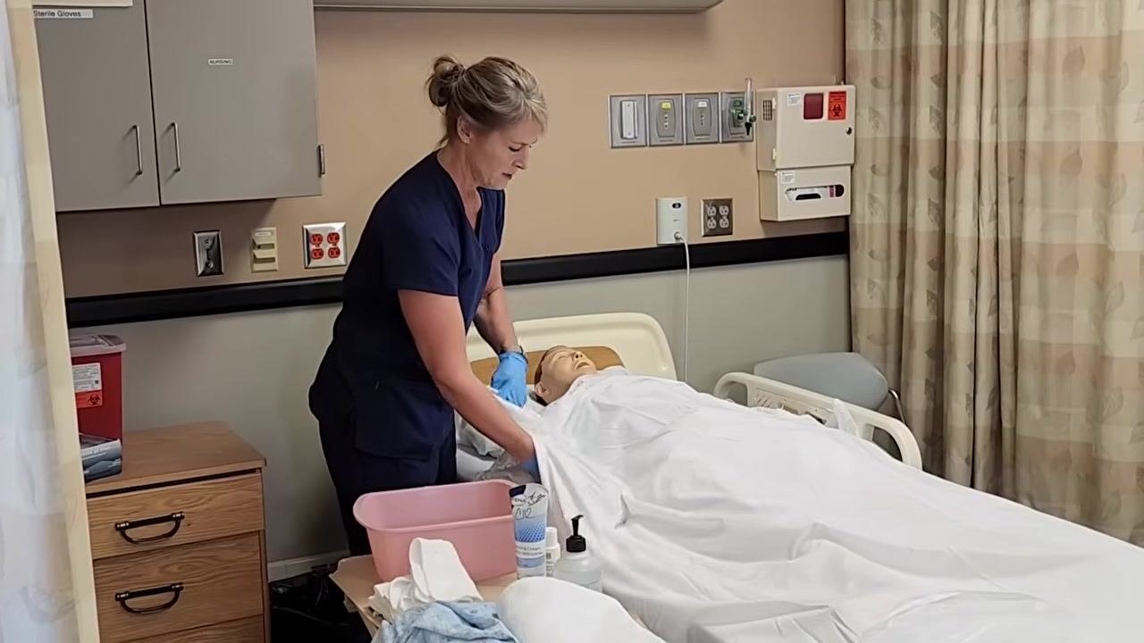 Nurse in blue scrubs placing a towel over the mannequin patient's chest, preparing for face washing. Supplies and a pink basin are on the bedside table.