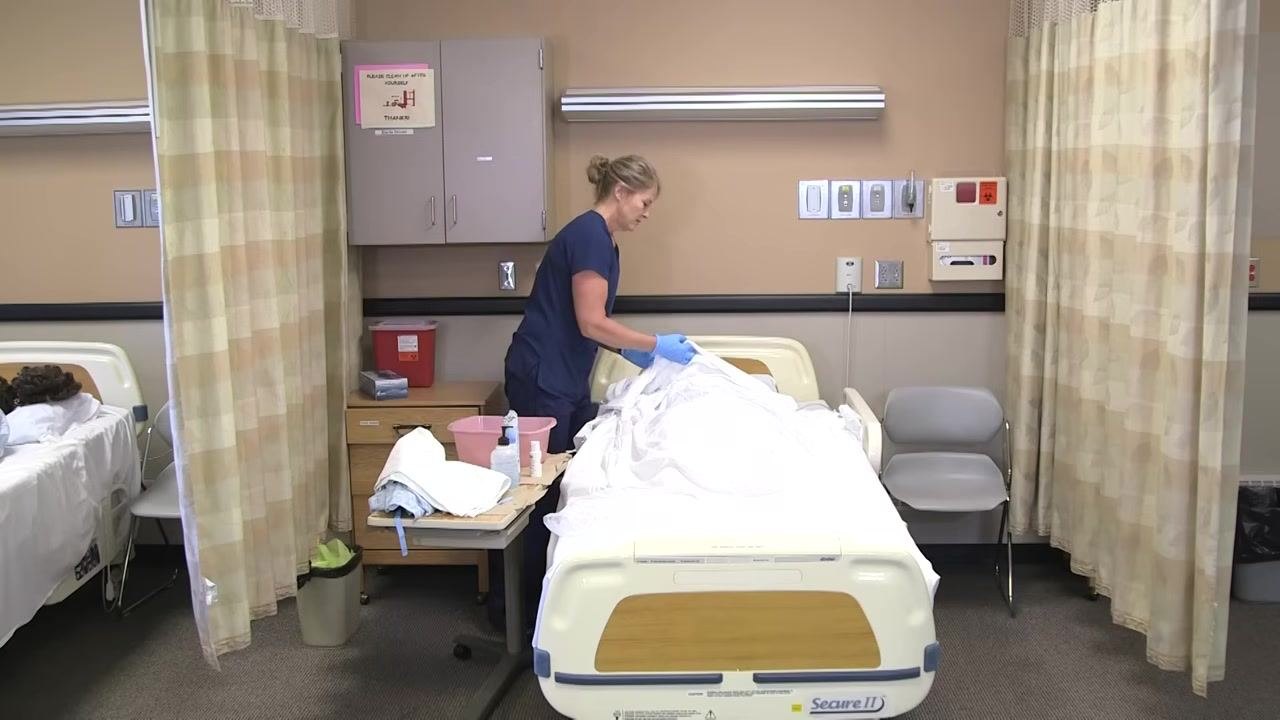 Nurse in blue scrubs gently patting the mannequin patient's face dry with a towel.