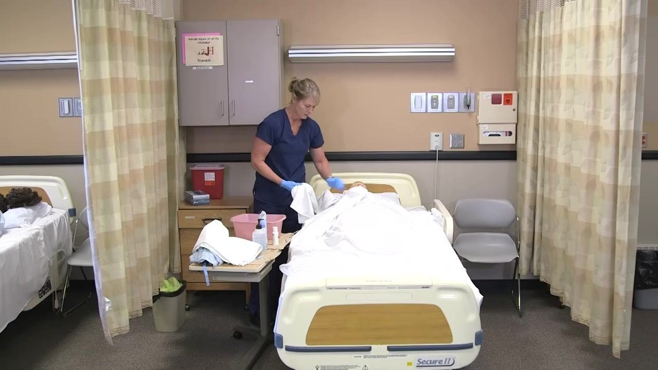 Nurse in blue scrubs placing a towel under the mannequin patient's arm, preparing for arm washing.