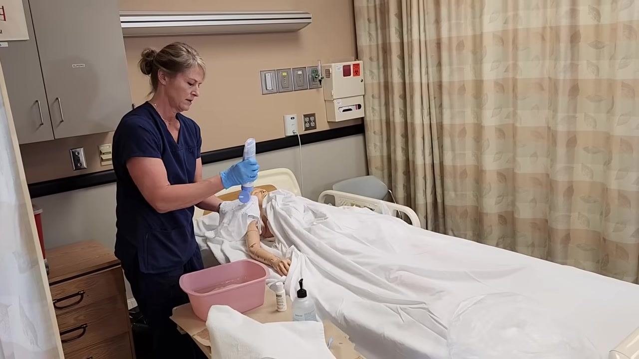 Nurse in blue scrubs removing the gown from the mannequin patient's arm while keeping them covered with a sheet.