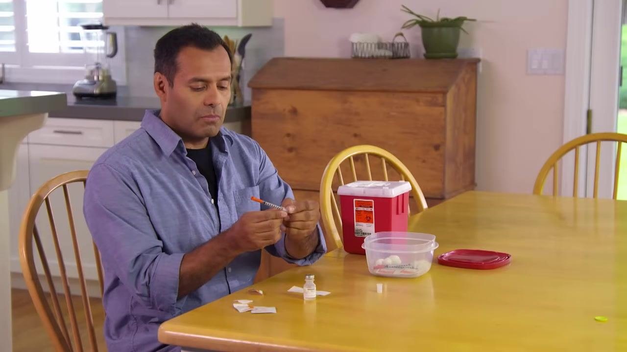 A person inserting the needle of a syringe into the top of a medication vial on a kitchen table.