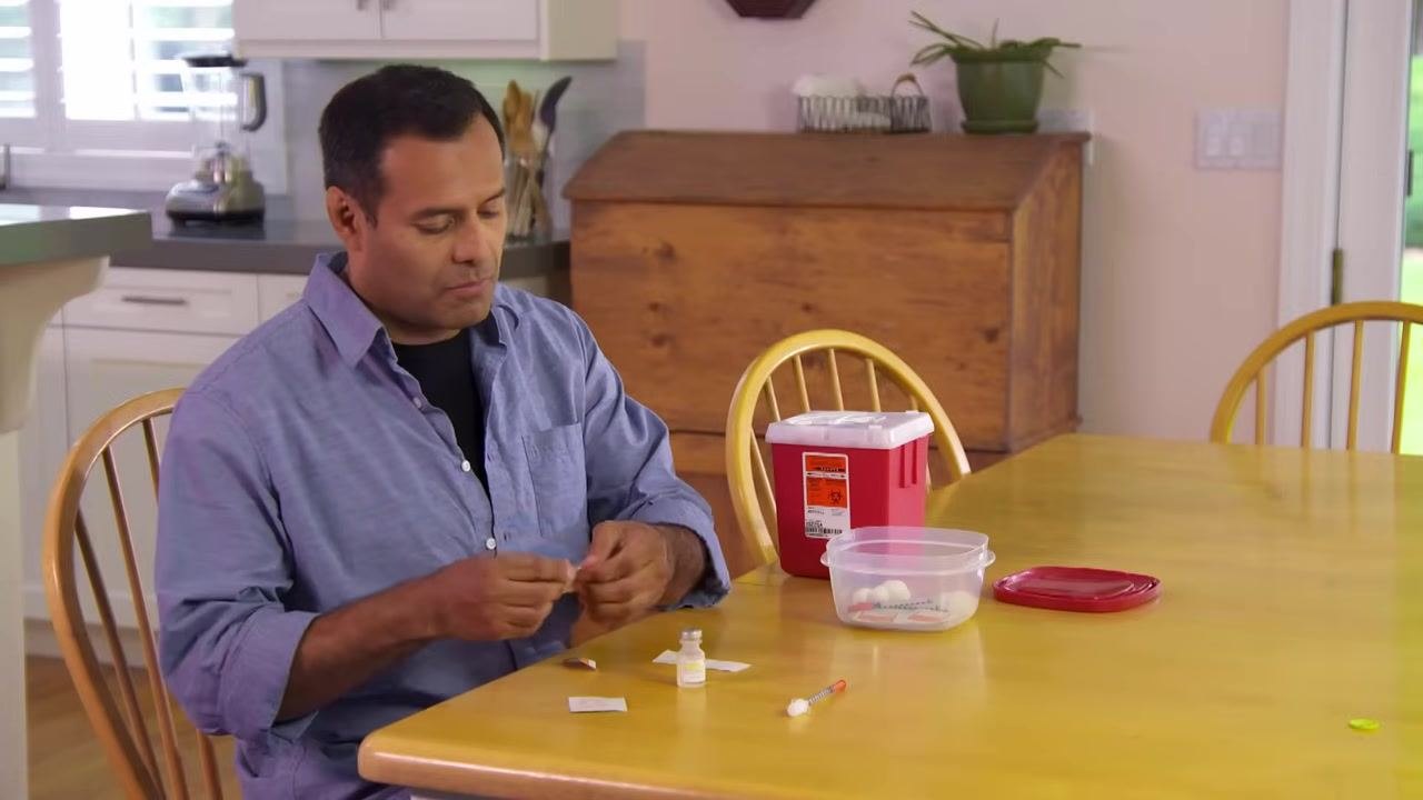 A person removing the cap from a syringe plunger at a kitchen table, with a vial, alcohol pads, and a sharps container visible.