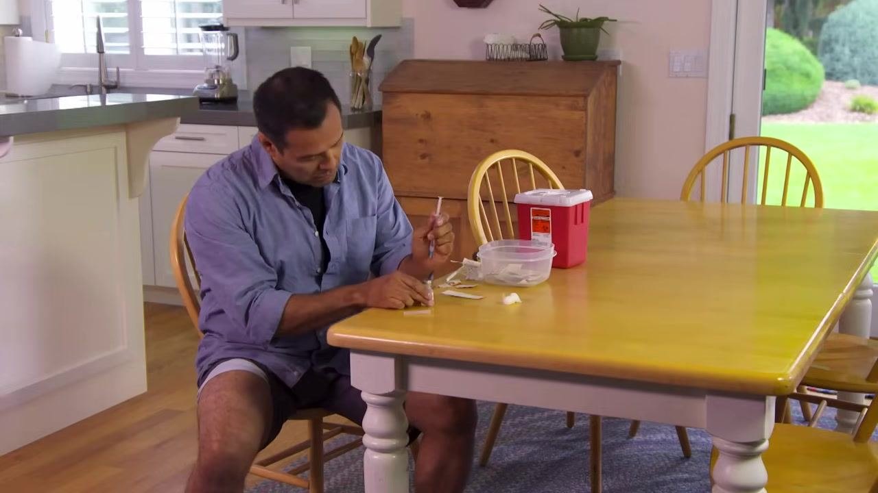 A person inserting a syringe needle into a vial at a kitchen table, with supplies and a sharps container nearby.