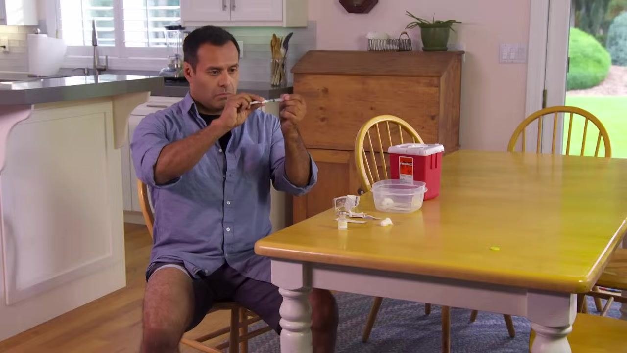 A person screwing a needle onto a syringe at a kitchen table, with supplies and a sharps container nearby.