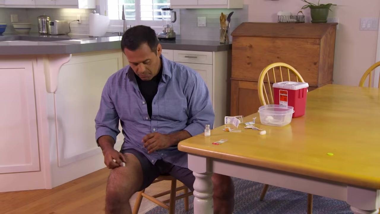 A person placing a used needle into a sharps container on a kitchen table, with other medical supplies nearby.
